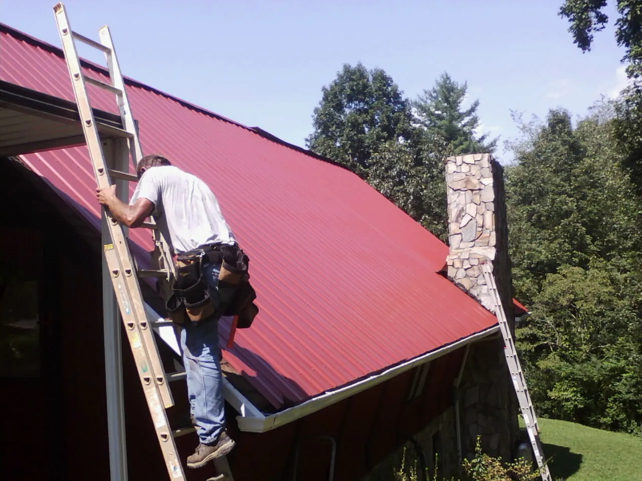 Roofer on a ladder beside a red metal roof and stone chimney — Soffit & Fascia Repair by Roofing Brothers in Jim Thorpe